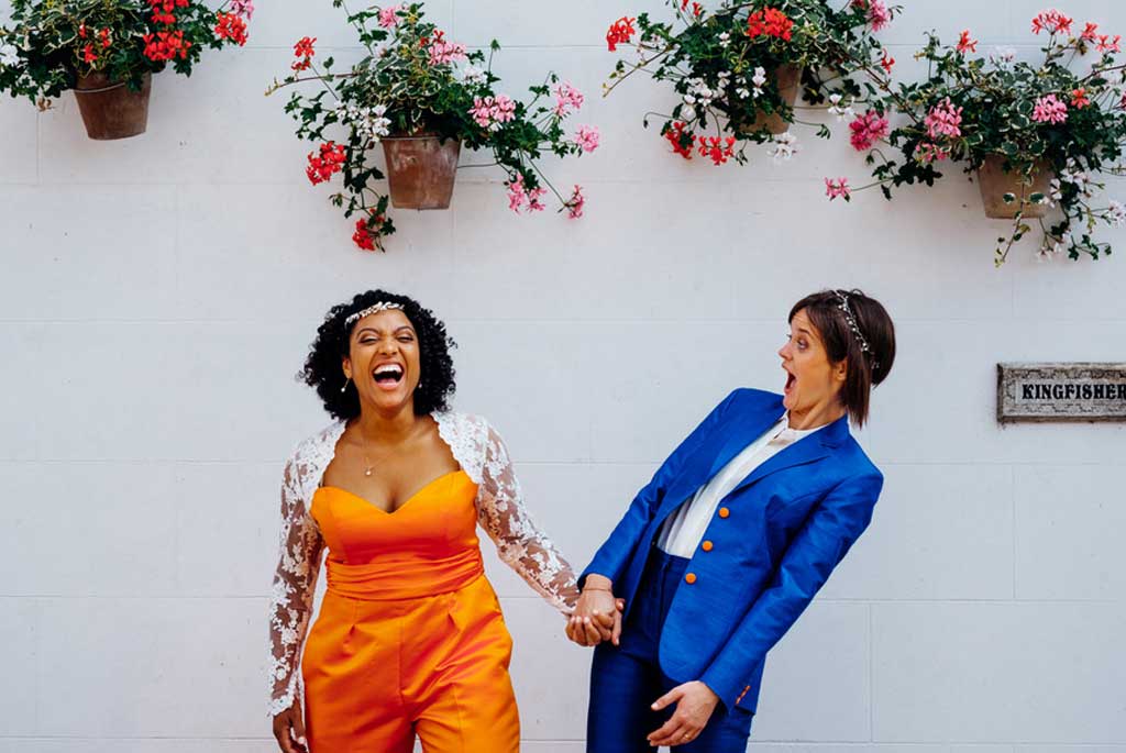 Two happy brides laughing together on their wedding day, both wearing colorful tailored suits; one orange and one blue. The pair tied the knot in the UK with unique womens wedding rings