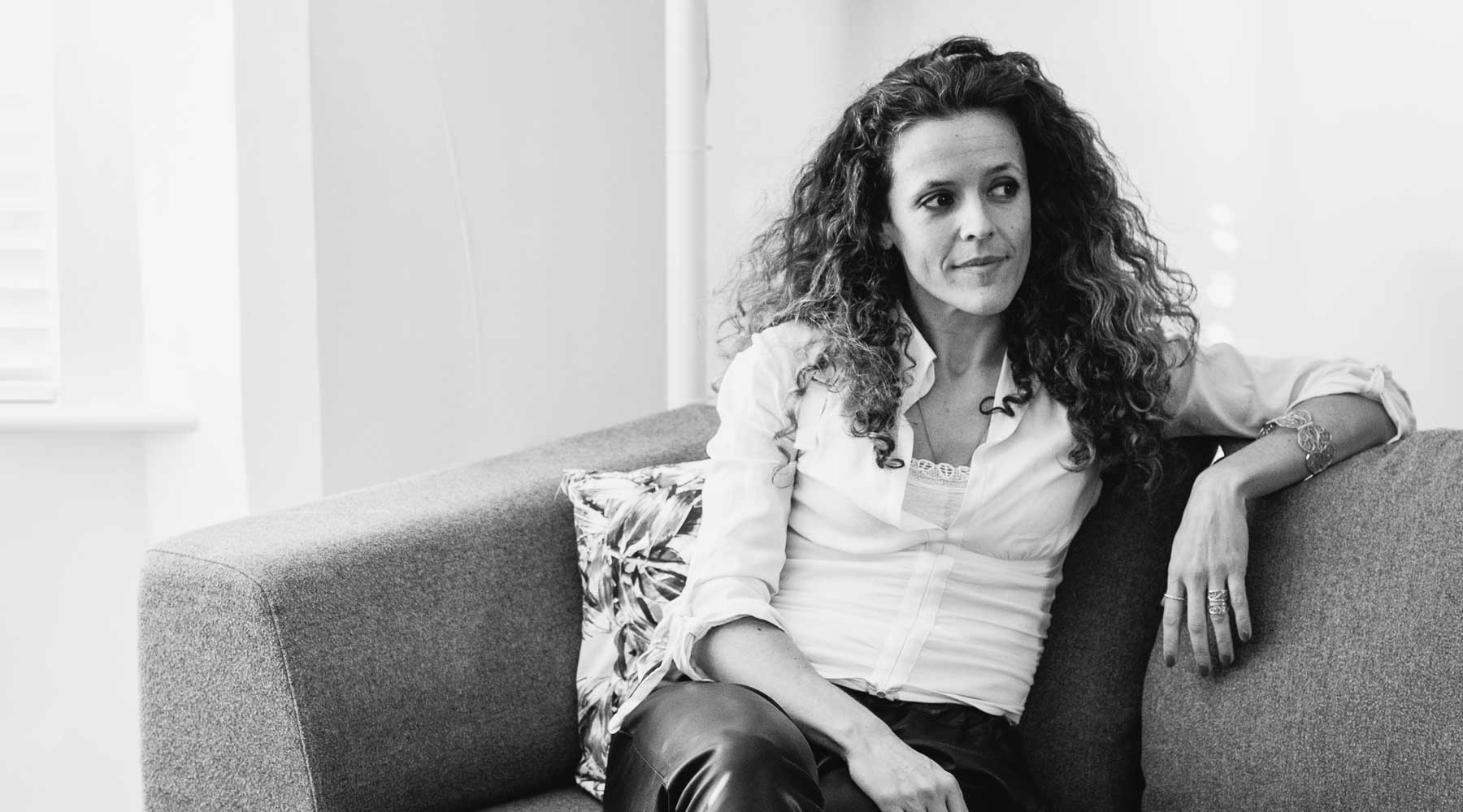 Black and white portrait of award-winning bespoke jewellery designer Arabel Lebrusan. Sitting on a sofa, she wears a white button-down shirt, dark leather trousers, and looks off to the left