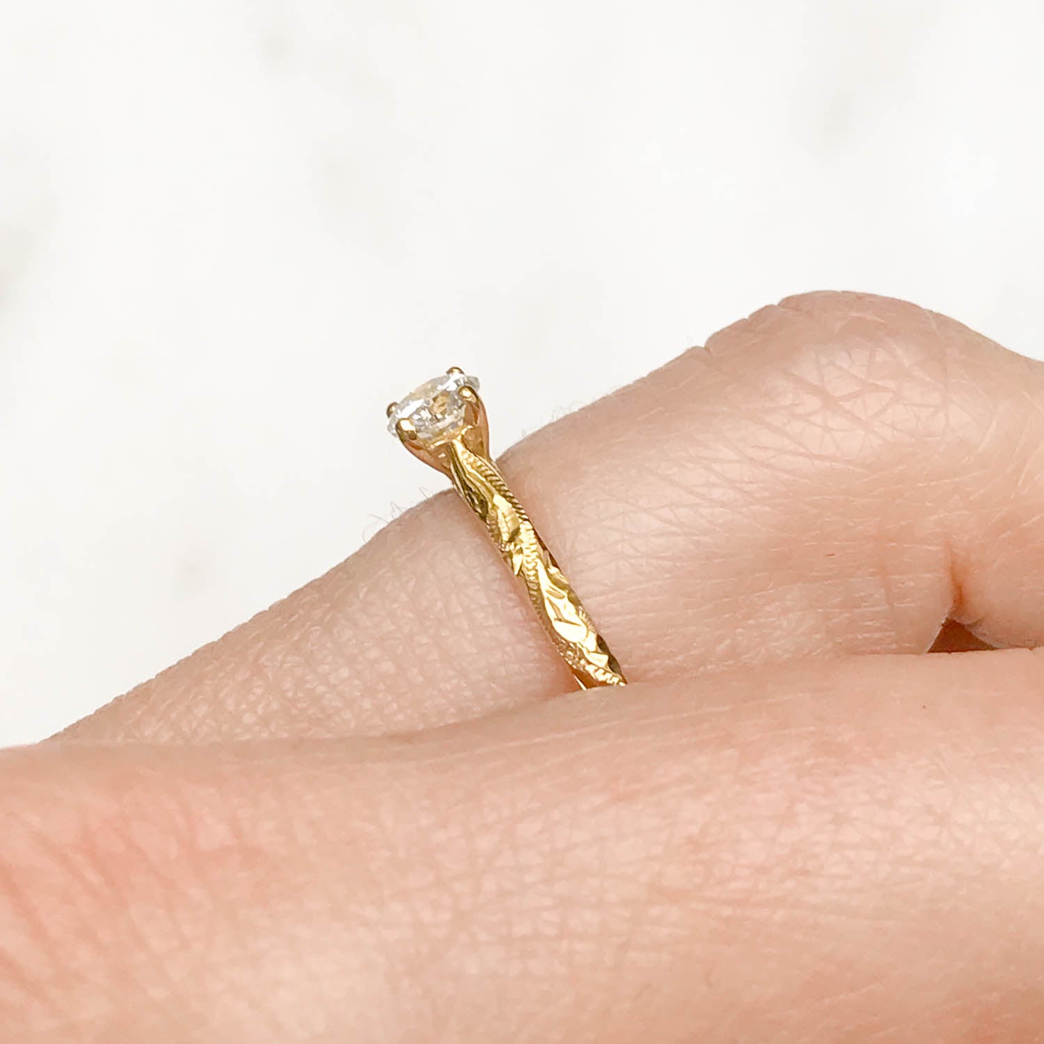 Solitaire engagement ring with a slim 18ct yellow gold band hand-engraved with intricate scrolls, and a 0.33ct round brilliant-cut diamond in a traditional four-claw setting, on hand side view to display scroll engraving