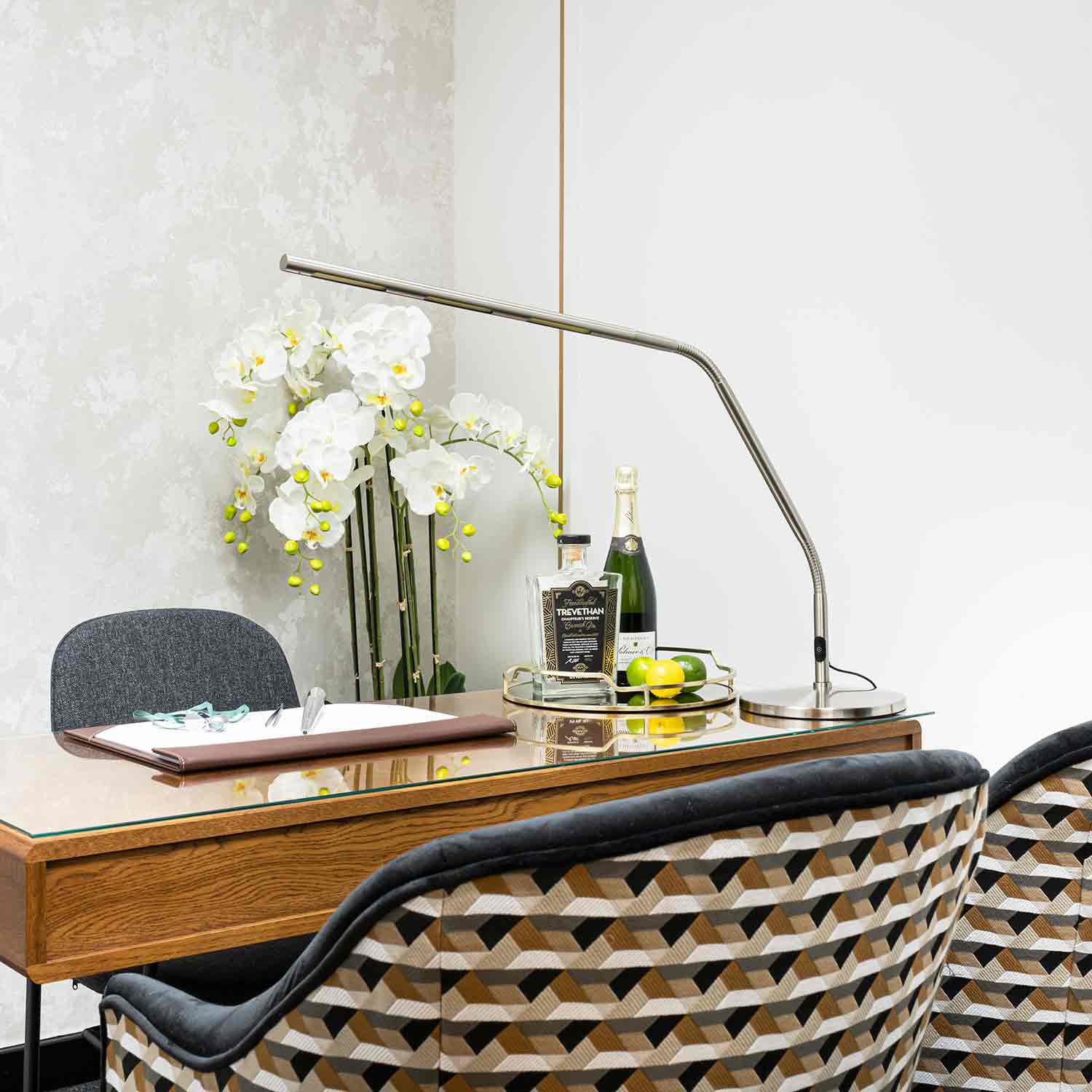 A London designer jewellery consultation room at the London Diamond Bourse. Two chairs upholstered in geometric fabric sit on one side of a polished wooden desk. A spray of white orchid-like flowers and liquor bottles decorate the desk