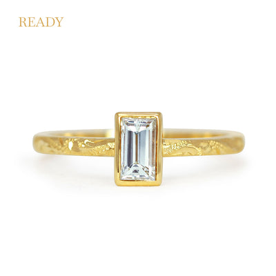 Lebrusan Studio Fancy Hera ready to wear engagement ring with a recycled baguette solitaire diamond in a rub-over setting; an 18ct recycled gold band is hand engraved with scrolls