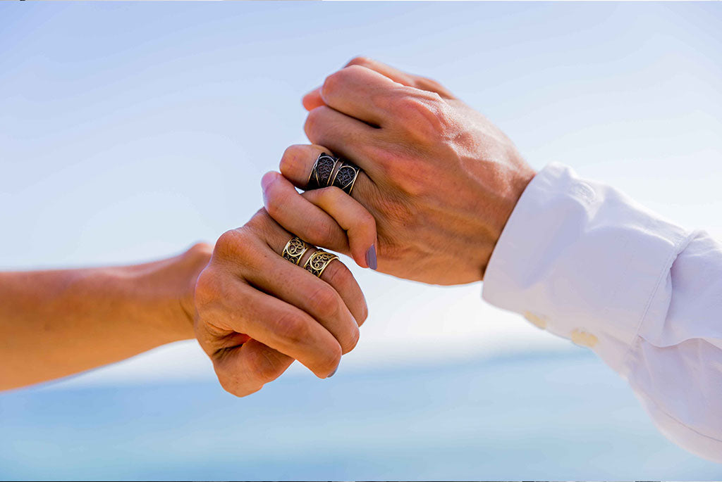 Two hands linking fingers in a sign of promise; both wearing commitment rings. The unique gold bands are hand-crafted aesthetic wedding rings, unique and custom made