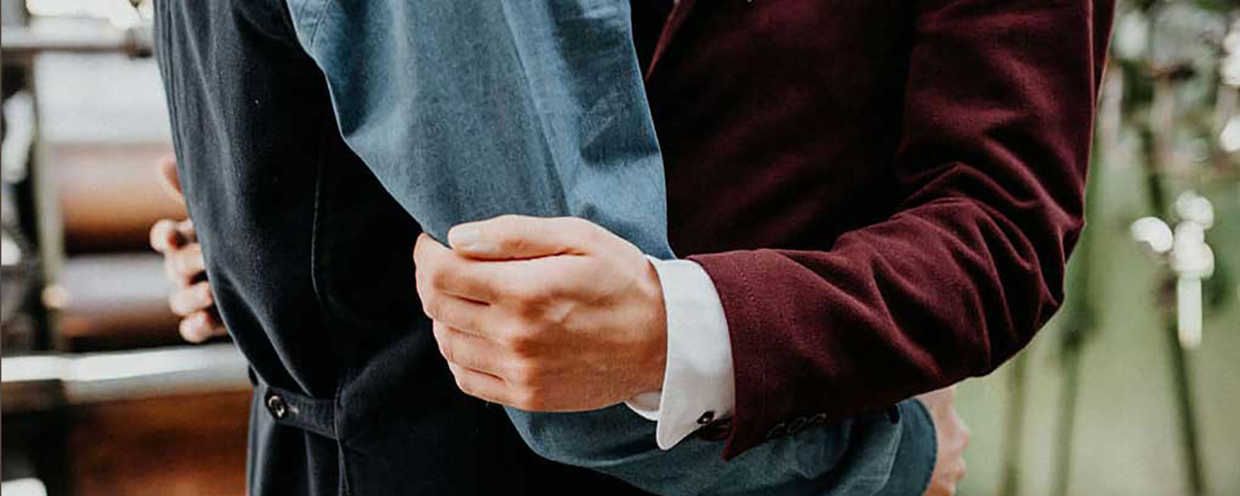 A faceless shot of two torsos wearing fitted suits and embracing, presumably a couple of white gay men at their wedding