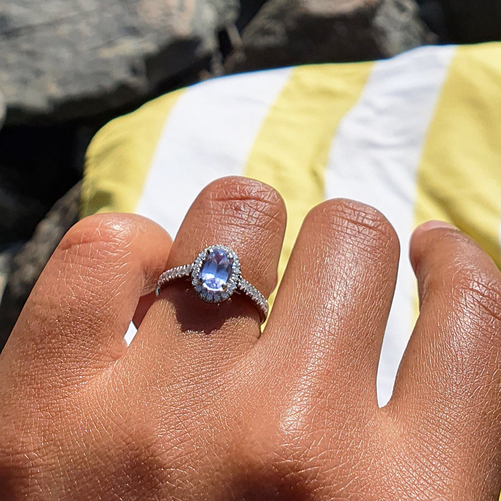 A custom diamond and sapphire engagement ring on a client's hand in the sunshine. Centre-stage is a lilac oval-cut fair-traded sapphire, secured within a halo of small conflict-free diamonds. The shoulders are also set with diamonds. Ethical jewelry UK