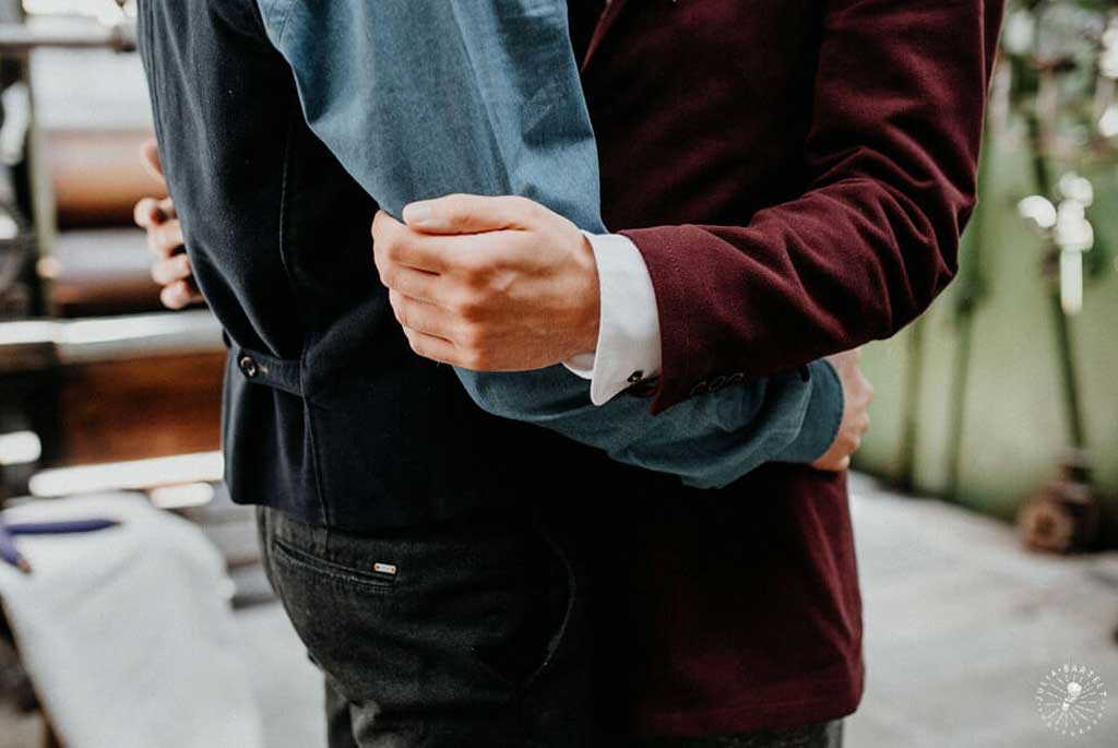 Two grooms on their wedding day, embracing each other in their suits. Both wear mens designer wedding bands, custom made and cast in ethical gold