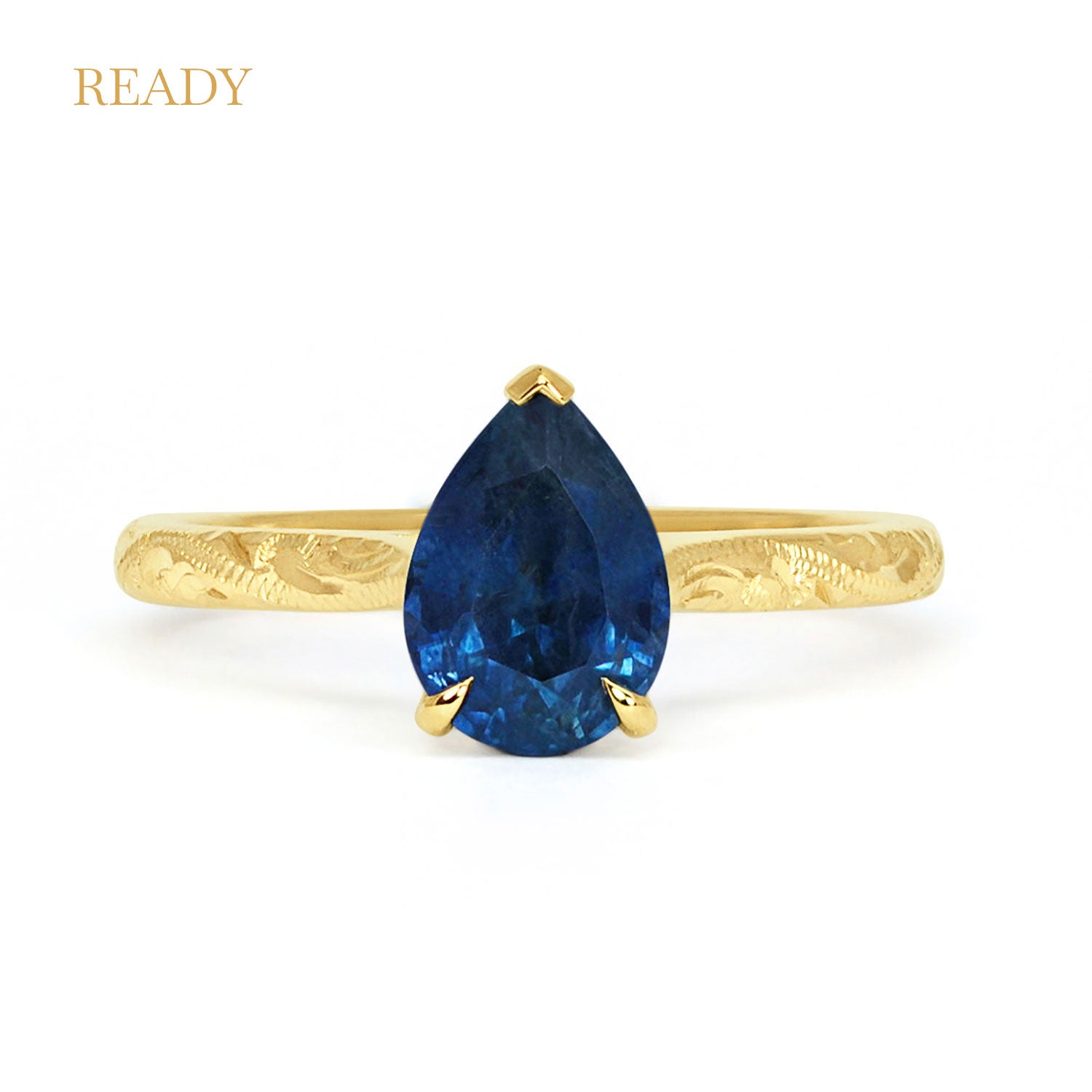 Solitaire engagement ring with a large pear-cut blue sapphire secured in a classic claw solitaire setting. 18ct recycled gold is hand engraved with scrolls