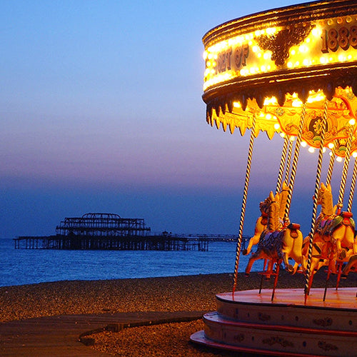Brighton's burnt-out West Pier at sunset, the sky and sea vibrant shades of blue, violet and pink. A colourful and illuminated fairground carousel is in the foreground