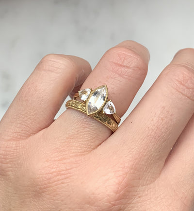 A D-shaped 2mm wedding band in 18ct recycled yellow gold, carefully hand-engraved with a repetitive orange blossom motif and a border of milgrain beading, paired with a one-off trilogy engagement ring set with white marquise sapphires